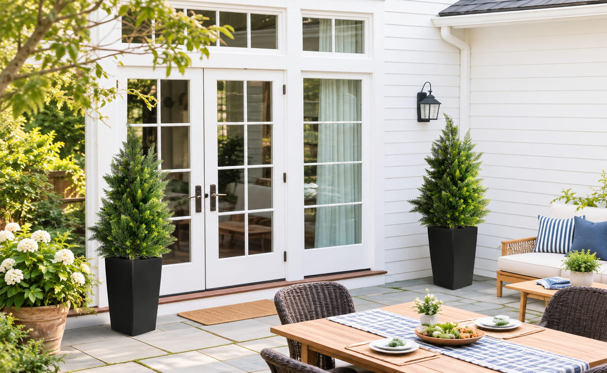 Two artificial cedar trees in black planters on a patio.