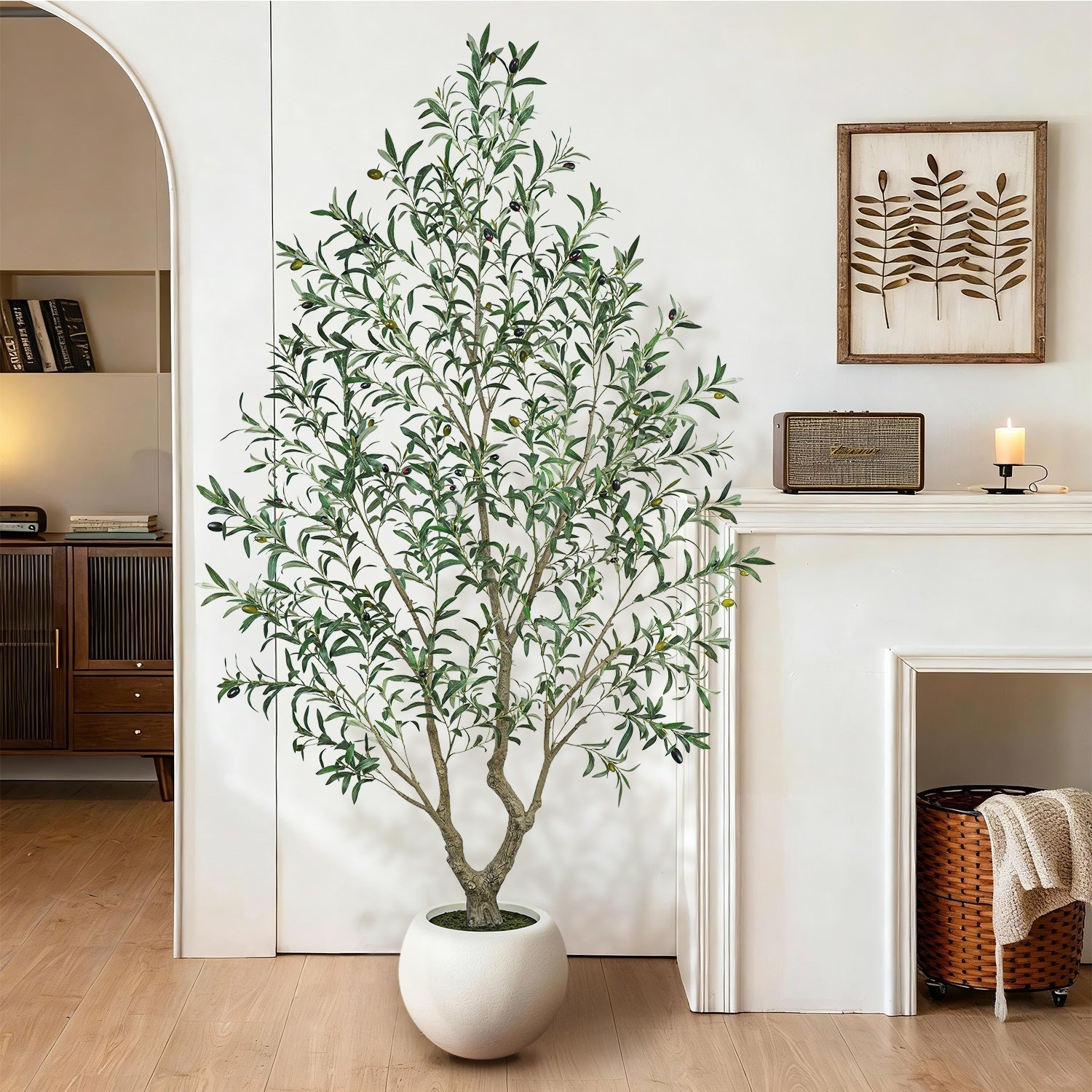 Large realistic artificial olive tree in a white round sphere pot placed in a modern cozy living room next to a fireplace