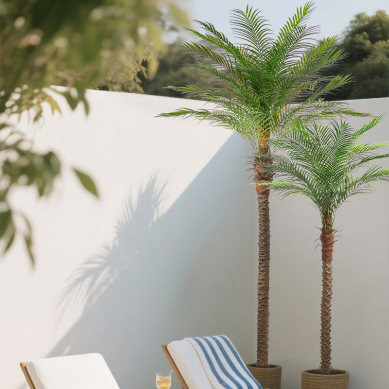 Two artificial palm trees in pots against a white wall with a chair and book in the foreground.